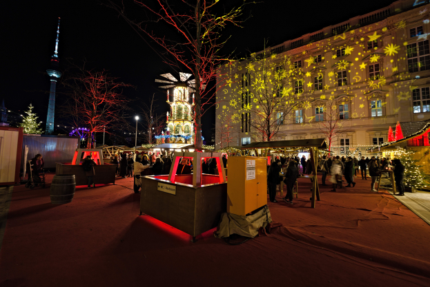 Ein geschäftiger Weihnachtsmarkt in Berlin, Deutschland, mit Menschen um beleuchtete, festliche Stände, Bäume, Gebäude, Laternenpfähle und einen Turm im Hintergrund unter einem dunklen Himmel.