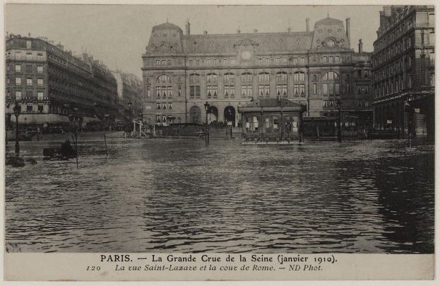 Schwarz-weiß-Foto einer überfluteten Straße in Paris mit Gebäuden, Straßenlaternen, Lichtern, Fahrzeugen und einem Himmel im Hintergrund, mit Text unten.