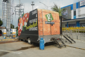 Ein großer Lastwagen parkt vor einem Gebäude mit einer Tafel, auf der "Teilen Sie ein positiv überladenes Erlebnis" steht, mit Stufen, einem Zylinder auf dem Boden, Geländern, Strommasten mit Drähten, Laternenmasten, Bäumen und einer bewölkten Himmel im Hintergrund.