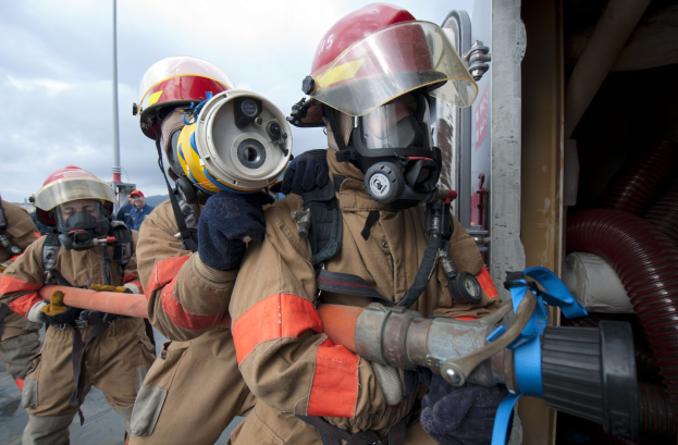 Feuerwehrleute in Schutzausrüstung, einer hält einen Schlauch, mit einem Mast und bewölktem Himmel im Hintergrund und Rohren auf der rechten Seite.