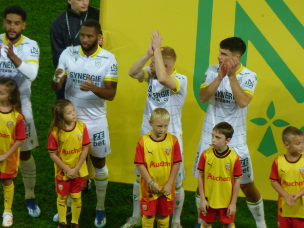 Eine Gruppe junger Menschen in Fußballtrikots steht auf einem Fußballfeld und klatscht feiernd in die Hände mit einer 'Ligue 1'-Platte im Hintergrund.