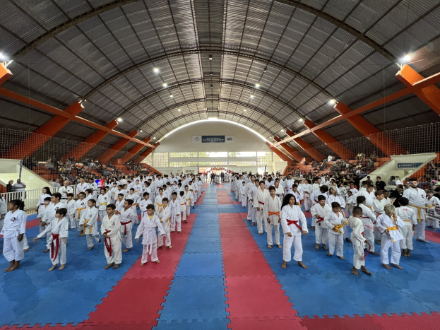 Große Gruppe von Menschen auf einer blauen und roten Matte in einer Turnhalle mit Zuschauern auf Treppen und Deckenbeleuchtung, Hintergrund zeigt Taekwondo-Veranstaltungsplakate.