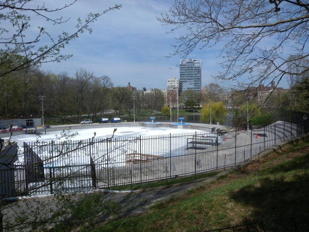 Eissporthalle in einem Park umgeben von einem Metallzaun, Gras, Bäumen, Fahrzeugen, Straßenlaternen, einem Gewässer, Gebäuden und einem bewölkten Himmel.