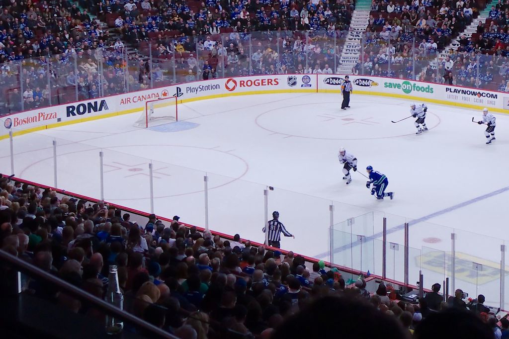 Vier Männer spielen Hockey auf einem Eisplatz, mit zwei in der Nähe des Tors und des Zauns, Zuschauern auf Stühlen im Vorder- und Hintergrund sowie einer Treppe im Abstand.