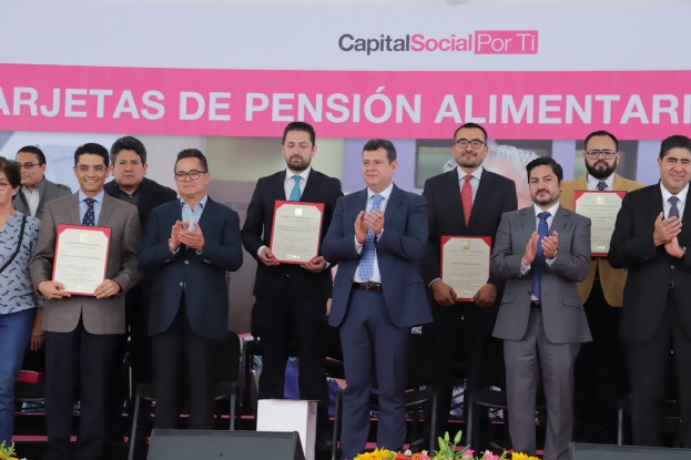 Eine Gruppe von Menschen auf einer Bühne, die klatschen und Zertifikate halten, mit Stühlen in einem Halbkreis darum herum und einem Banner mit der Aufschrift "Capital Social for Ti" im Hintergrund, zusammen mit Blumensträußen und Rednern unten.