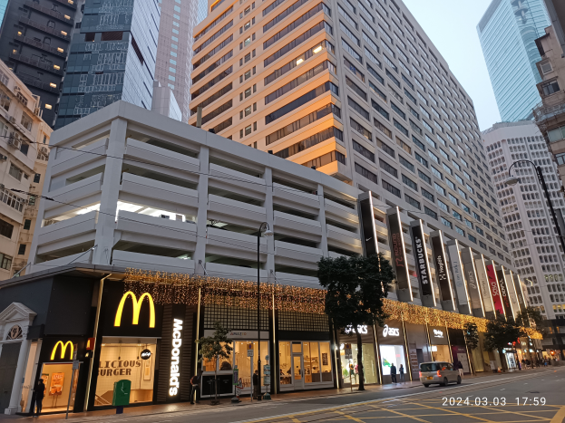 Ein McDonald's-Restaurant auf einer belebten Stadtstraße, umgeben von hohen Gebäuden, Straßenlaternen, Verkehrspoller, fahrenden Fahrzeugen, Fußgängern, Bäumen und einem klaren blauen Himmel.