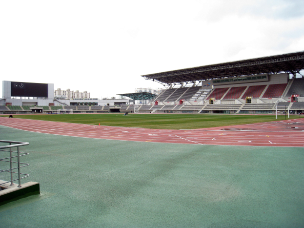 Ein großes Stadion mit einer Laufbahn, einem Geländer auf der linken Seite, Gebäuden im Hintergrund und einem klaren blauen Himmel.