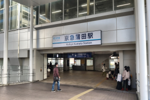 Menschen mit Taschen durch eine Bahnhofshalle gehen, eine zentrale Tafel mit Text, Säulen, Geländer, Glaswände und Deckenlampen; ein Mann in Fußbekleidung ist in der Mitte zu sehen.