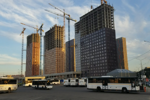 Eine Gruppe von Bussen, die auf einem Parkplatz neben höhen Gebäuden geparkt sind, mit Straßeninfrastruktur und Bäumen unter einem bewölktem Himmel.
