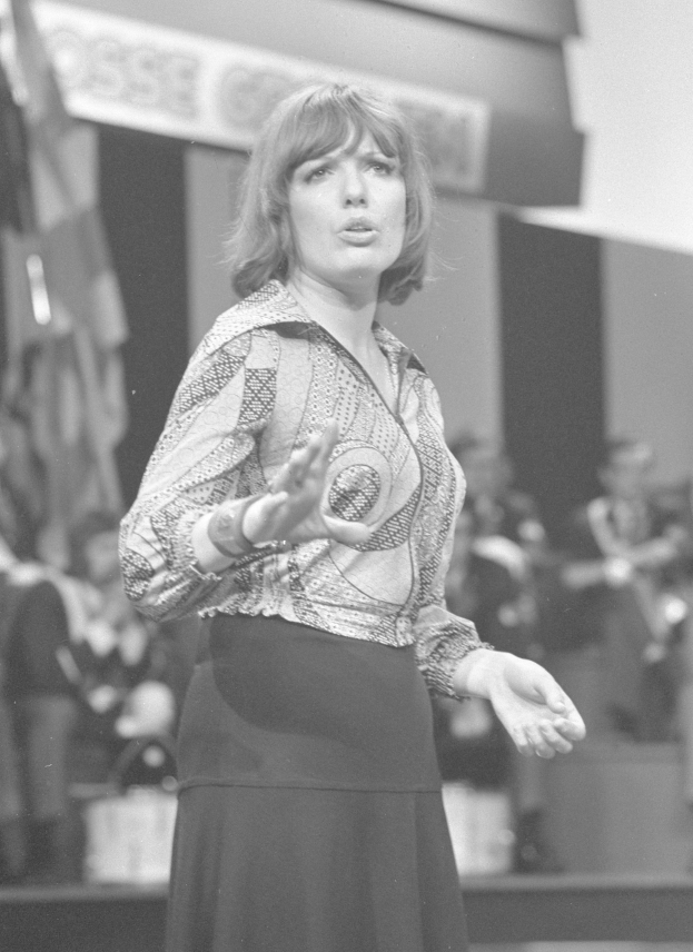 Eine Frau in einem Schwarz-Weiß-Foto steht auf einer Bühne umgeben von einer Gruppe von Menschen, trägt ein weißes Hemd und einen schwarzen Rock und hat ihr Haar zu einem Dutt gebunden und einen ernsten Ausdruck.