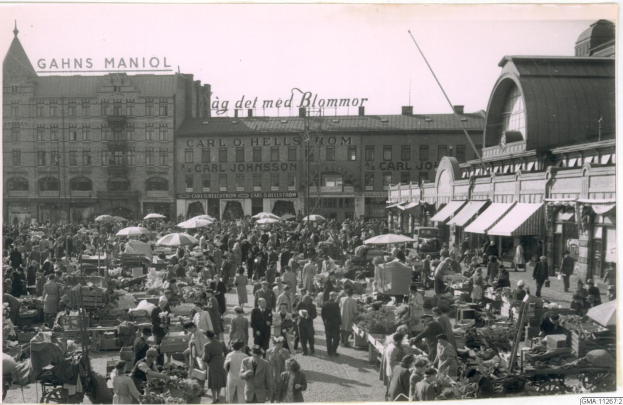 Schwarz-weißes Foto eines belebten Berliner Markts mit Menschen, Gemüsewagen und Gebäuden im Hintergrund.
