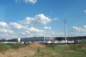 Mercedes-Benz-Fabrik in Deutschland mit Fahrzeugen auf der Straße, Grünflächen, Industriegebäuden und einem bewölkten Himmel.