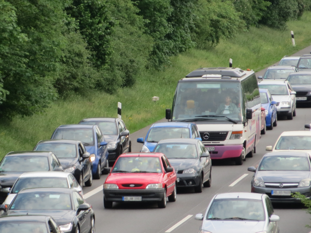 Ein Stau auf einer Autobahn mit vielen Autos und einem Lieferwagen, Menschen sind in den Fahrzeugen zu sehen und im Hintergrund gibt es Bäume und Gras.