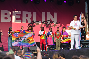 Gruppe von Menschen auf einer Bühne mit einem "Berlin as Fuck"-Plakat mit Lautsprechern, Mikrofonen und einer feiernden Menge darunter
