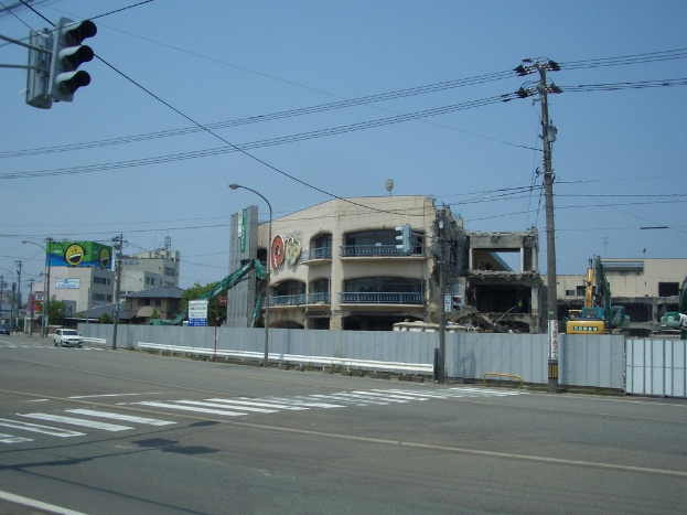 Eine Stadtstraße mit Fahrzeugen, Strommasten, Laternen, einer Ampel auf der linken Seite, Gebäuden, Bäumen und dem Himmel im Hintergrund und einem Gebäude in derForeground, das abgerissen wird.