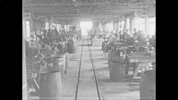 Schwarzes und weißes Foto einer Fabrik mit Arbeitern, die Maschinen bedienen, nummerierte Fässer, eine Eisenbahnschiene, Säulen und Deckenleuchten während der industriellen Revolution.
