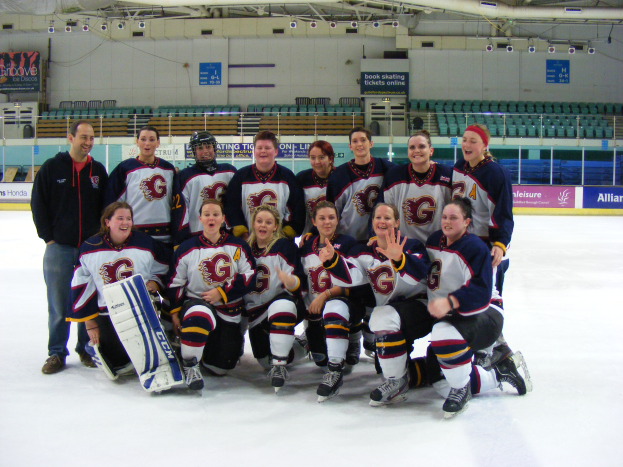 Eine Gruppe von Frauen-Eishockeyspielerinnen in Helmen und Schlittschuhen posiert für ein Foto auf dem Eis, wobei eine einen Hockey-Schläger hält, Stühle, Tafeln mit Text, Geländer und Deckenleuchten im Hintergrund zu sehen sind.