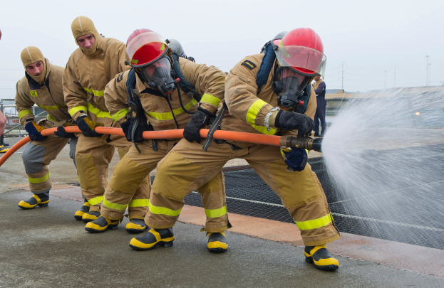 Feuerwehrleute in Schutzausrüstung nutzen einen Schlauch, um ein Feuer zu löschen, mit Strommasten und Kabeln im Hintergrund und einer Person mit einer Rohrleitung auf der linken Seite.