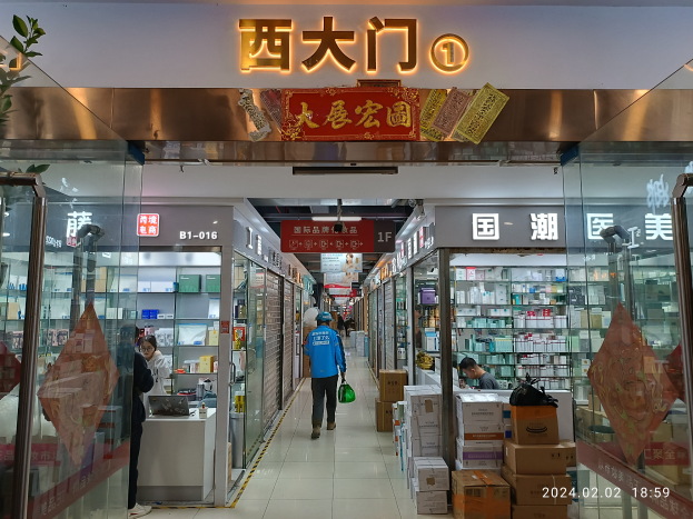 Das Innere einer chinesischen Apotheke mit Menschen, die hindurchgehen, Glaswänden auf beiden Seiten, einer Pflanze auf der linken Seite, Regalen, die mit verschiedenen Gegenständen gefüllt sind, Kartons und anderen Gegenständen, die auf dem Boden verstreut sind, und einem Namensschild mit Text oben.