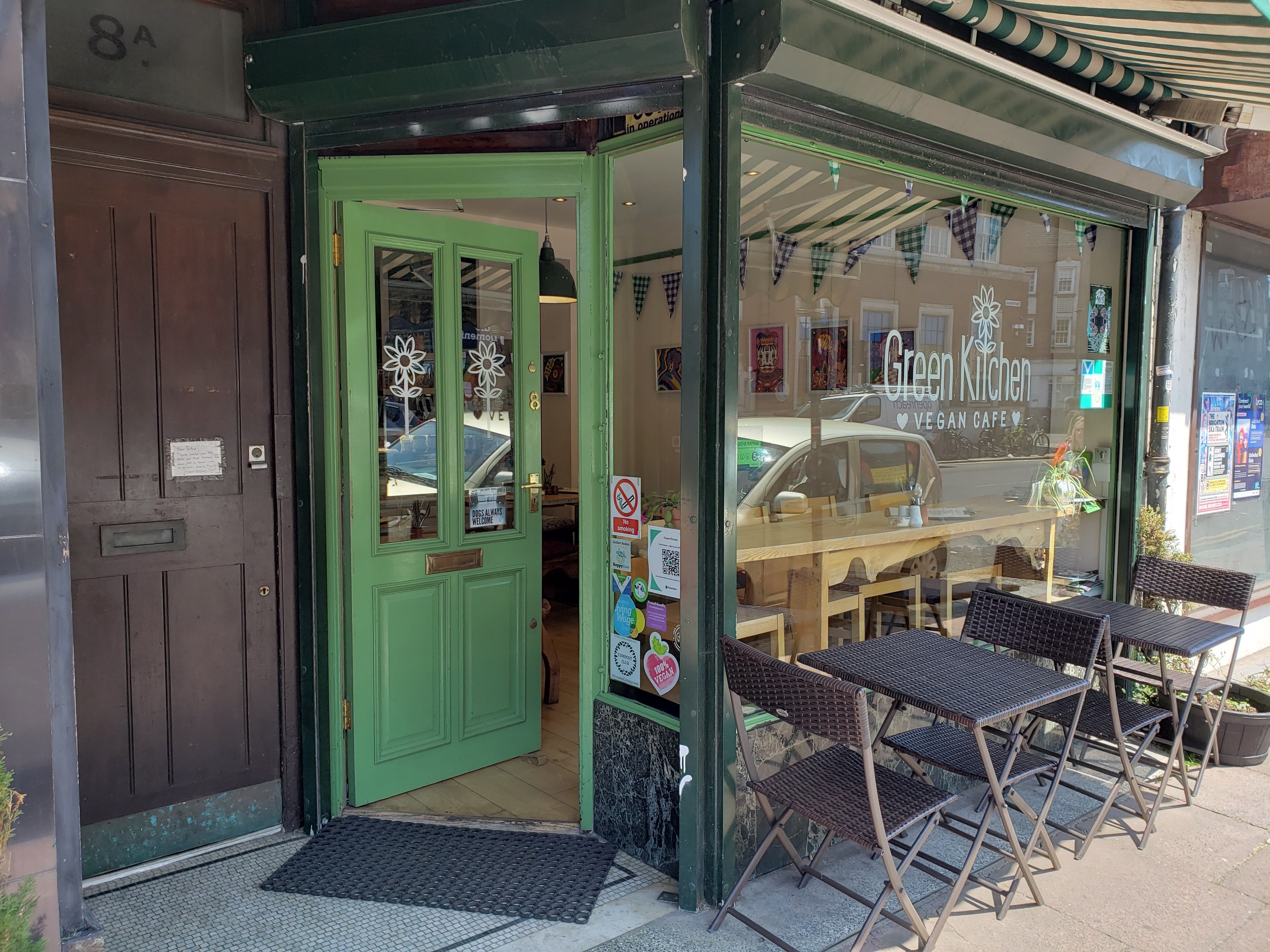 Außensitzbereich des Green Kitchen Vegan Cafe mit Tischen, Stühlen, Pflanzen, einer Fußmatte und einer Glastür mit Postern, der in einen Innenbereich mit Tischen, Stühlen und einer Deckenlampe führt.