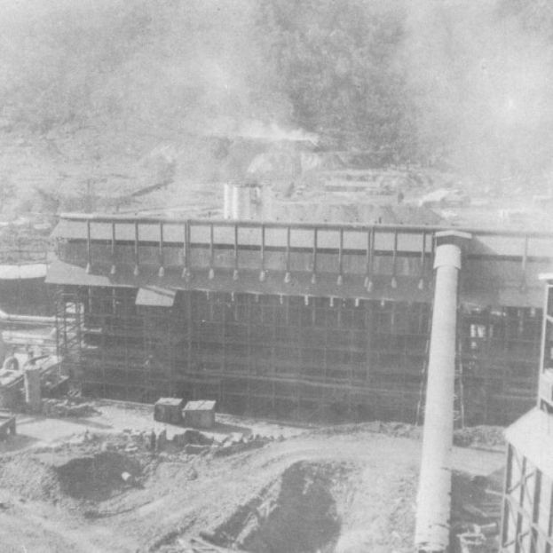 Ein Schwarz-Weiß-Foto einer Fabrik mit Rauch, der aus dem Schornstein aufsteigt, umgeben von Gebäuden, Pfählen und anderen Gegenständen am Boden.