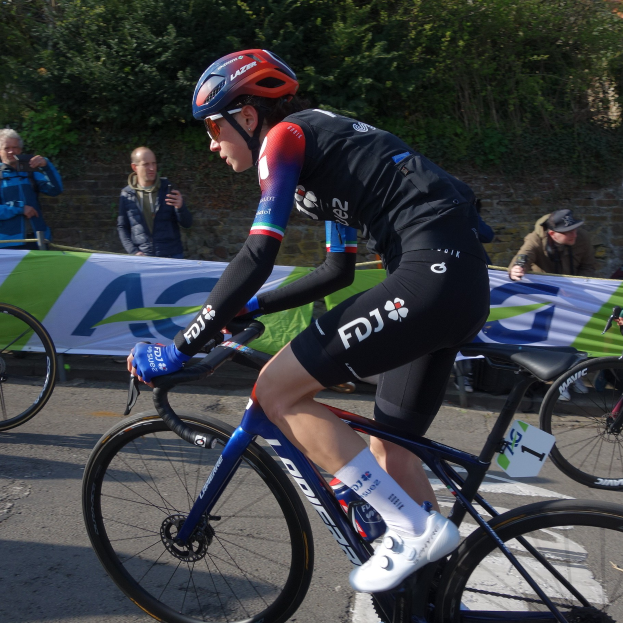 Professionelle Radsportlerin Katie Archibald fährt mit Helm, Brille, Handschuhen und Schuhen auf einem Fahrrad eine Straße entlang, umgeben von einer Menge und einem Banner.