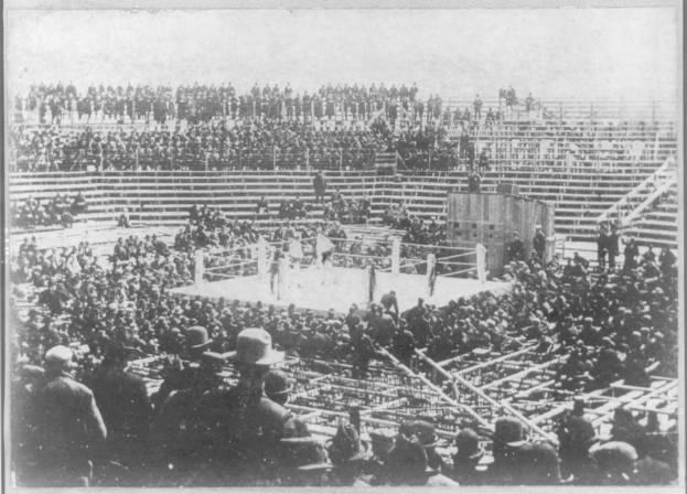 Ein Schwarz-Wei├č-Foto einer Menge in einem Stadion, mit einem Boxring und einigen Menschen drumherum in der Mitte.