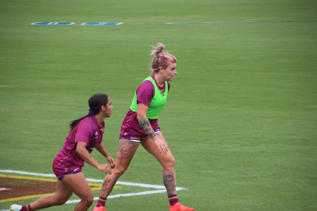 Zwei Frauen beim Fussballspielen auf einem Feld, eine trägt ein grünes T-Shirt und Shorts, die andere ein pinkes Kleid, beide haben Tätowierungen an Händen und Füssen.