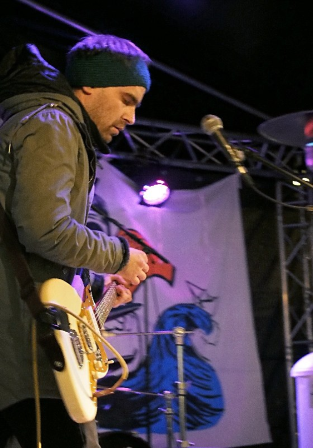 Ein Mann in einer Jacke und einer Mütze spielt eine E-Gitarre auf der Bühne mit einem Mikrofon davor, einem Schlagzeug rechts von ihm und Scheinwerfern und einem Banner im Hintergrund.
